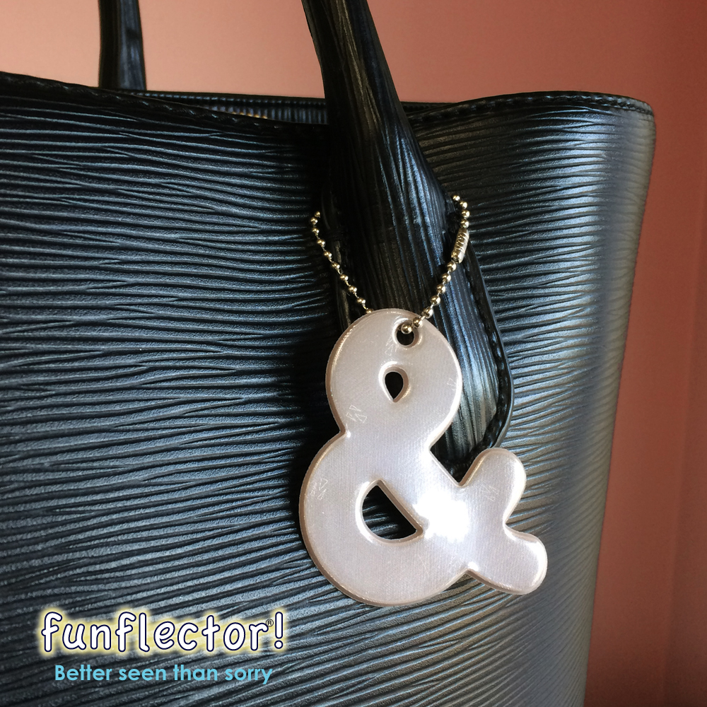 Ampersand safety reflector in sandalwood hanging on the handle of a bag.